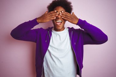 Mor kazak giyen genç Afro-Amerikalı adam izole edilmiş pembe arka planda duruyor. Gözleri neşeli ve komik gülümseyen ellerle örtüyor. Kör kavram.