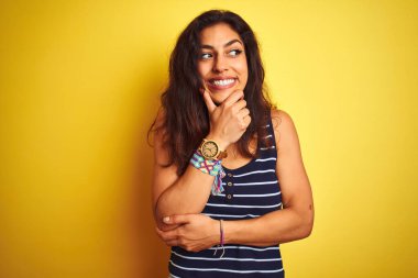 Çizgili tişört giyen genç ve güzel bir kadın izole edilmiş sarı arka planda duruyor ve elleri çenesinde soru sormayı düşünüyor, düşünceli bir ifade. Gülen ve düşünceli bir yüz. Şüphe kavramı.