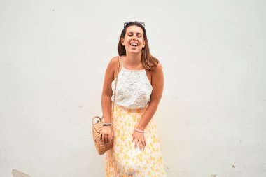 Güzel kız beyaz duvara yaslanıyor, genç arkadaş canlısı kadın mutlu bir yaz gününde gülümsüyor.