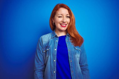 Genç, güzel, kızıl saçlı, kot gömlek giyen, mavi izole edilmiş arka plan üzerinde duran, kameraya seksi bir ifadeyle bakan, neşeli ve mutlu yüzlü bir kadın..