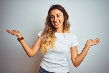 İzole edilmiş arka planda günlük beyaz tişört giyen genç ve güzel bir kadın gülümseyerek avuçlarını açıyor, sunuyor ve reklam karşılaştırması yapıyor ve dengeyi sağlıyor.