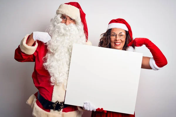 上了年纪的夫妇穿着圣诞老人的服装 拿着横幅挂在孤立的白色背景上 满脸惊讶地指着自己 — 图库照片