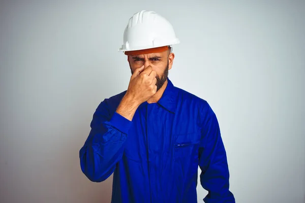 Handsome indian worker man wearing uniform and helmet over isolated white background smelling something stinky and disgusting, intolerable smell, holding breath with fingers on nose. Bad smells concept.