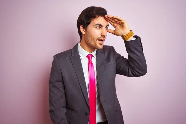 若いハンサムなビジネスマンがスーツを着て 孤立したピンクの背景の上に立ってネクタイは非常に幸せと遠くに手を頭の上で見て笑顔 検索の概念 — ストック写真
