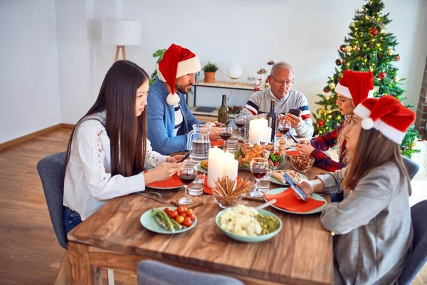 サンタの帽子をかぶっている美しい家族は笑顔で自信を持っています 自宅でクリスマスを祝うスマートフォンを使用してロースト七面鳥を食べる — ストック写真