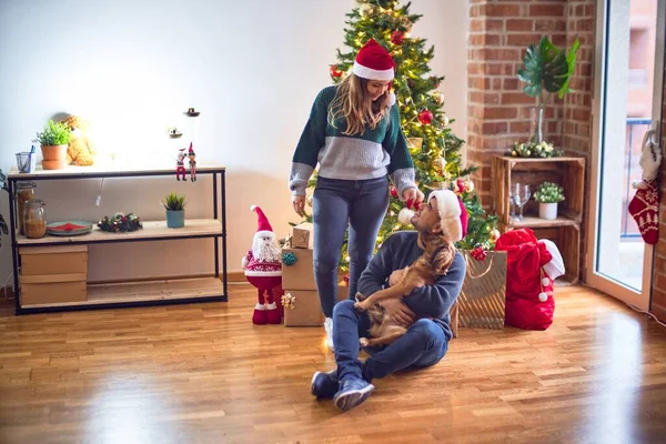 年轻漂亮的夫妇面带微笑 既快乐又自信 在家里 坐在地板上 头戴圣爪帽 抱着狗绕着圣诞树 — 图库照片