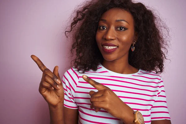 Young african american woman wearing striped t-shirt standing over isolated pink background smiling and looking at the camera pointing with two hands and fingers to the side.