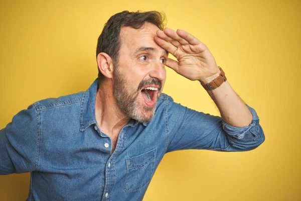 Handsome middle age senior man with grey hair over isolated yellow background very happy and smiling looking far away with hand over head. Searching concept.