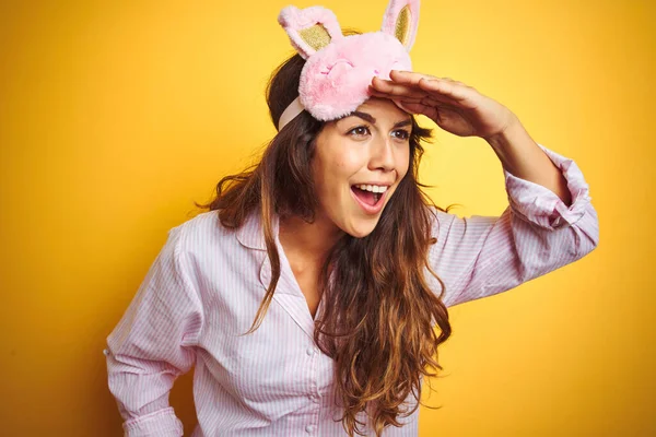 Young woman wearing pajama and sleep mask standing over yellow isolated background very happy and smiling looking far away with hand over head. Searching concept.