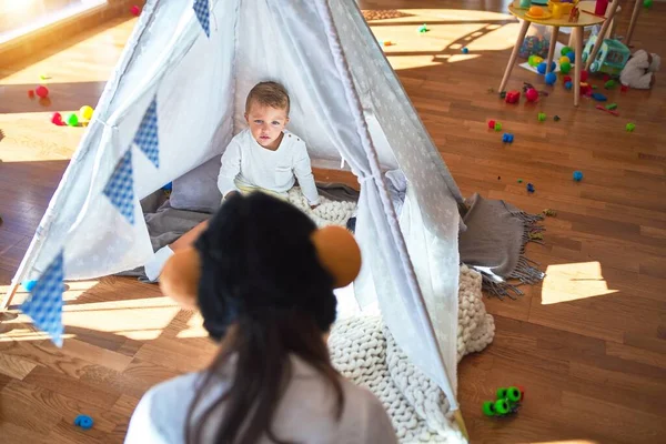 Beautiful teacher and toddler playing inside tipi around lots of toys at kindergarten