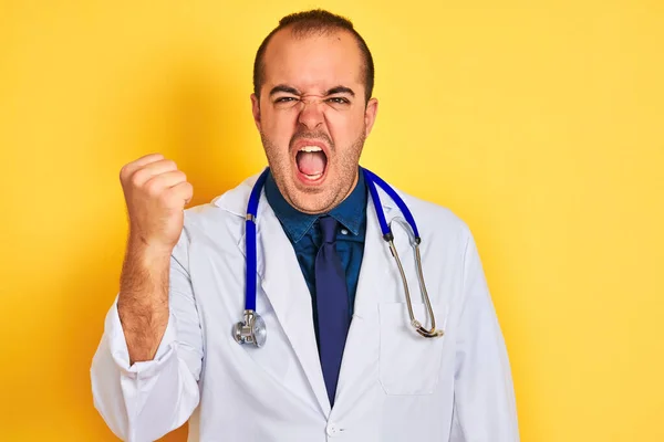 独立した黄色い背景の上にコートと聴診器を身に着けている若い医者の男は怒りで叫んでいる間に拳をフラストレーションと激怒を上げ怒っている 激怒と積極的な概念 — ストック写真