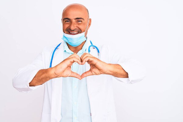 Middle age doctor man wearing stethoscope and mask over isolated white background smiling in love showing heart symbol and shape with hands. Romantic concept.