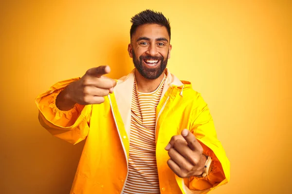 Young indian man wearing raincoat standing over isolated yellow background pointing fingers to camera with happy and funny face. Good energy and vibes.