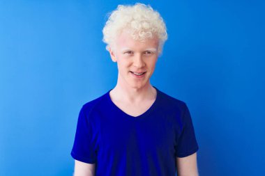 Sıradan tişörtler giyen genç albino bir adam mutlu ve havalı bir gülümsemeyle izole edilmiş mavi arka planda dikiliyor. Şanslı kişi..