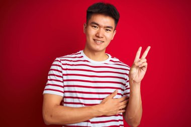 Çizgili tişört giyen Asyalı genç bir adam izole edilmiş kırmızı arka planda dikiliyor ve kameraya gülümsüyor zafer işareti yapıyor. İki numara..