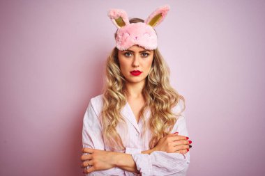 Pembe izole edilmiş arka plan üzerinde pijama ve uyku maskesi takan genç ve güzel bir kadın şüpheci ve gergin, yüzünde çapraz kollu tasvip etmeyen bir ifade var. Negatif kişi.