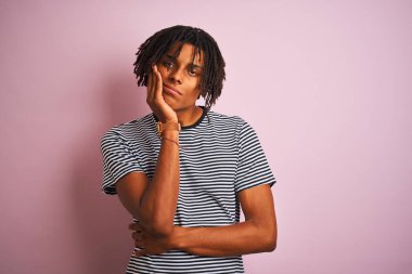 Rastalı tişörtlü Afro bir adam izole edilmiş pembe arka planda dikilirken kolları çaprazlanmış depresyondan sıkılmış ve yorgun görünüyor..