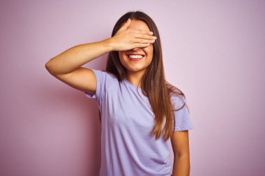 İzole edilmiş pembe arka plan üzerinde sıradan tişörtler giyen genç ve güzel bir kadın. Gülümsüyor ve elleri yüzünde şaşkınlıktan gözleri kapatıyordu. Kör kavram.