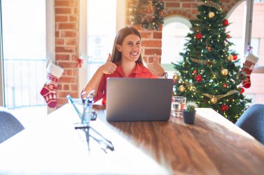 Güzel bir kadın evde dizüstü bilgisayarla çalışıyor Noel ağacı civarında el hareketleriyle olumlu jest yapıyor, gülümsüyor ve mutlu. Neşeli ifade ve kazanan jest.