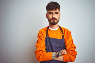 İzole edilmiş beyaz arka plan üzerinde duran önlüklü genç bir esnaf şüpheci ve gergin, yüzünde çapraz kollu tasvip etmeyen bir ifade. Negatif kişi.
