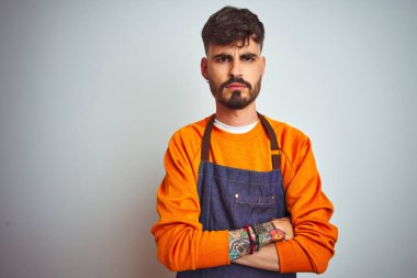 Young shopkeeper man with tattoo wearing apron standing over isolated white background skeptic and nervous, disapproving expression on face with crossed arms. Negative person. clipart