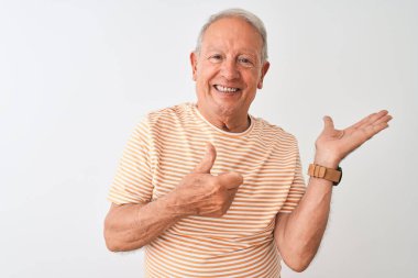 Kıdemli, gri saçlı, çizgili tişört giyen, izole edilmiş beyaz arka planda duran, avuç içini gösteren ve baş parmaklarıyla hareket eden, mutlu ve neşeli gülümseyen bir adam.