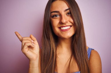 İzole edilmiş pembe arka planda duran tişört giyen genç ve güzel bir kadın mutlu bir şekilde elini ve parmağını kenarı işaret ediyor.