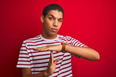 İzole edilmiş kırmızı arka plan üzerinde çizgili tişört giyen genç yakışıklı Arap adam elleri ve ciddi yüzüyle jest yapıyor.