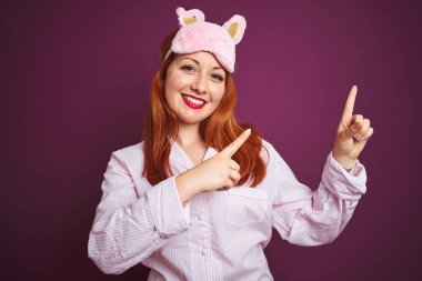 Genç, güzel, kızıl saçlı, pijama giymiş ve maskeli bir kadın. Mor, izole bir arka plan. Gülümsüyor ve kameraya bakıyor. İki eliyle ve parmaklarıyla işaret ediyor..
