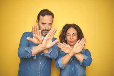 Güzel orta yaşlı çift, izole edilmiş sarı arka planda dikiliyorlar. Reddedilme ifadesi, kollar ve avuç içleri kesişiyor.