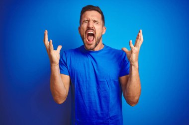 Mavi izole edilmiş arka plan üzerine gündelik tişört giyen genç yakışıklı adam çılgın ve başarıyı kollarını kaldırarak ve gözlerini kapayarak heyecanla bağırarak kutluyor. Kazanan konsept