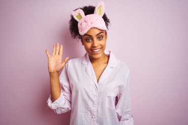 Genç Afro-Amerikalı kadın pijama giyip maske takıyor pembe arka planda. Mutlu ve güler yüzlü, dostça bir jest yapıyor.