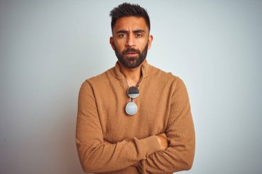 Zarif süveter ve güneş gözlüğü takan Hintli bir adam izole edilmiş beyaz arka planda şüpheli ve gergin duruyor. Yüzünde çapraz kollu tasvip etmeyen bir ifade var. Negatif kişi.