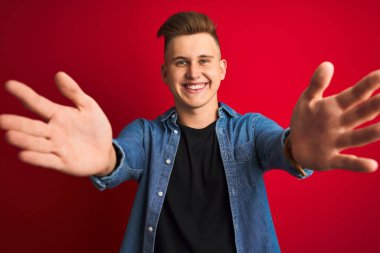 İzole edilmiş kırmızı arka plan üzerinde duran kot gömlek giyen genç yakışıklı adam kucaklanmak için kollarını açmış gülümseyen kameraya bakıyor. Mutluluğu kucaklayan neşeli bir ifade..