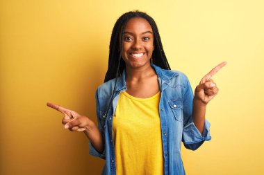 İzole edilmiş sarı arka planda duran kot gömlekli genç Afro-Amerikan kadın parmaklarıyla farklı yönleri gösteren kendine güvenle gülümsüyor. Reklam için boşluk kopyala