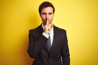 Takım elbiseli ve kravatlı genç yakışıklı bir işadamı izole edilmiş sarı arka planda dikiliyor ve parmakları dudaklarında sessiz olmak istiyor. Sessizlik ve gizli konsept.