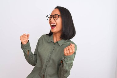 Yeşil tişörtlü ve gözlüklü genç Çinli bir kadın izole edilmiş beyaz arka planda çok mutlu ve heyecanlı bir şekilde kollarını kaldırıp, gülümseyerek ve başarı çığlıkları atarak zafer jesti yapıyor. Kutlama konsepti.