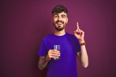 Dövmeli genç adam izole edilmiş mor arka planda duran su bardağını içerken bir fikir ya da soru işareti ile şaşırdı.