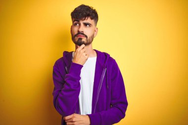 Dövmeli genç bir adam, mor spor kazak giyiyor, izole edilmiş sarı arka planda, elleri çenesinde soru sorarken, düşünceli bir ifade takınıyor. Düşünceli bir yüzle gülümsüyor. Şüphe kavramı.