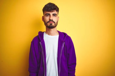 İzole edilmiş sarı arka plan üzerine mor kazak giyen dövmeli genç adam yüzünde ciddi bir ifadeyle rahatlamış. Basit ve doğal bir şekilde kameraya bakıyorsun..