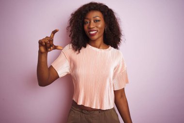 Genç Afro-Amerikalı kadın pembe arka plan üzerinde duran tişört giyiyor gülümsüyor ve el hareketleriyle küçük boy işareti yapıyor ve kameraya bakıyor. Ölçü kavramı.