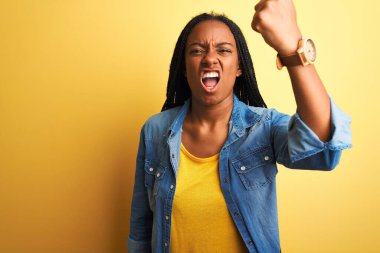 Genç Afro-Amerikalı kadın kot gömlek giyerek izole edilmiş sarı arka plan üzerinde duruyor kızgın ve kızgın yumruğunu kaldırıyor ve öfkeyle bağırıyor. Öfke ve agresif kavram.