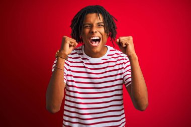 Kolları havada ve gözleri kapalı zaferi kutlayan gülümsemeyle başarı için heyecanla izole edilmiş kırmızı arka planda duran çizgili tişörtlü Afrikalı genç bir adam. Kazanan konsept.