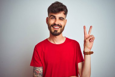 Kırmızı tişörtlü genç adam izole edilmiş beyaz arka planda dikiliyor. Kameraya gülümsüyor ve zafer işareti yapıyor. İki numara..