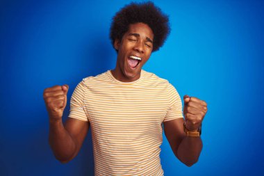 İzole edilmiş mavi arka plan üzerinde çizgili sarı t-shirt giyen Amerikalı bir adam çok mutlu ve heyecanlı bir şekilde kollarını kaldırıp, gülümseyerek ve başarı için çığlık atarak zafer hareketi yapıyor. Kutlama konsepti.