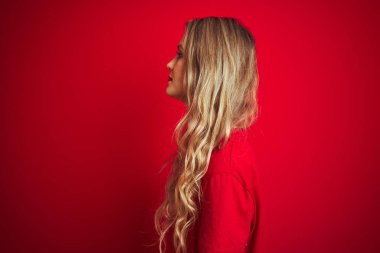 Temel tişört giyen genç bir kadın kırmızı izole edilmiş arka plan üzerinde duruyor yan tarafa bakıyor, rahat bir poz veriyor kendine güvenen bir gülümsemeyle doğal bir yüz ifadesi sergiliyor..