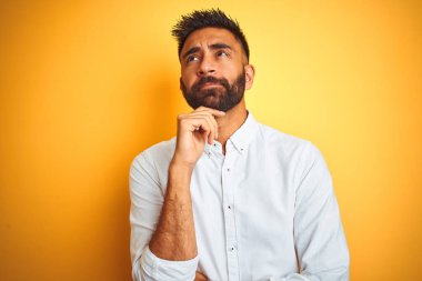 Hintli iş adamı, sarı arka plan üzerinde duran beyaz şık gömlekli, elleri çenesinde soru soran, düşünceli bir ifade takınıyor. Düşünceli bir yüzle gülümsüyor. Şüphe kavramı.