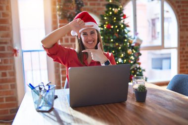 Noel 'de Noel Baba şapkası takıp dizüstü bilgisayarla çalışan güzel bir kadın gülümseyerek elleri ve parmaklarıyla mutlu bir yüzle çerçeve yapıyor. Yaratıcılık ve fotoğrafçılık kavramı.