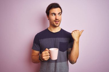 Dövmeli genç yakışıklı adam izole edilmiş pembe arka planda bir fincan kahve içiyor ve gülümseyen bir gülümsemeyle baş parmağıyla onu işaret ediyor.
