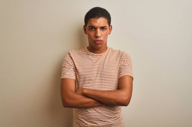 İzole edilmiş beyaz arka plan üzerinde çizgili tişört giyen genç yakışıklı Arap adam şüpheci ve gergin, yüzünde çapraz kollu tasvip etmeyen bir ifade var. Negatif kişi.